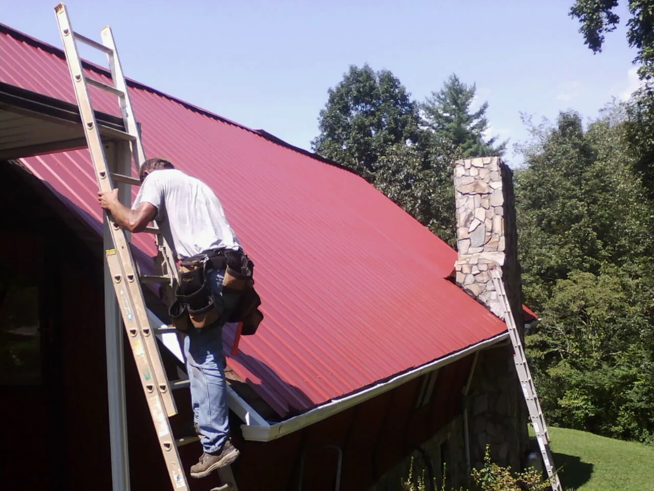 Roofer on a ladder beside a red metal roof and stone chimney — Soffit & Fascia Repair by Roofing Brothers in Bloomingburg